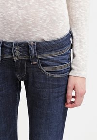 Los jeans de mezclilla azul oscuro tienen una cintura de altura media, cierre de dos botones y múltiples bolsillos con costuras en contraste. Textura suave y diseño ajustado.
