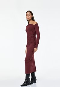 Eine Frau steht in einem langen, burgunderroten, gerafften Kleid mit einem schulterfreien Träger und schwarzen Absatzstiefeln vor einem schlichten weißen Hintergrund.