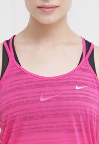 Top atletico rosa con spalline sottili, linee orizzontali sottili e un piccolo logo Nike bianco sul petto. Canottiera nera visibile sotto.