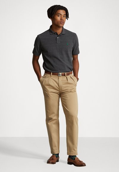 Polo de color gris oscuro con un logo verde, combinado con pantalones beige con pliegues y zapatos marrones. Los accesorios incluyen calcetines de rayas y un cinturón marrón.