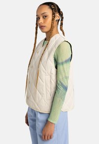 Gilet sans manches matelassé en crème, avec un motif en losanges, des boutons-pression et des poches latérales. Porté sur un haut vert texturé à manches longues.