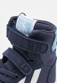 Mörkblå högskurna sneakers i mocka och tygmaterial. Har en blå accent, kardborreband och vita ränder längs sidan.