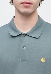 Γκρι πόλο μπλούζα με υφή υφάσματος. Διαθέτει λαιμόκοψη με τρία κουμπιά και μικρό χρυσό λογότυπο στο αριστερό στήθος.