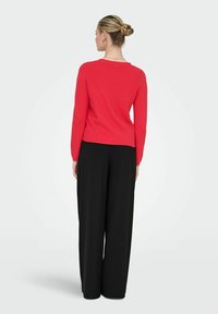 Femme avec chignon tressé, portant un haut rouge à manches longues et un pantalon noir à jambes larges, debout de dos sur un fond uni.