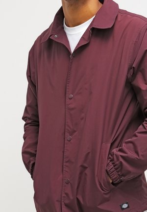 Mann trägt eine bordeauxrote leichte Jacke mit Druckknöpfen und Seitentaschen über einem weißen Rundhalsshirt, Hände in den Taschen.