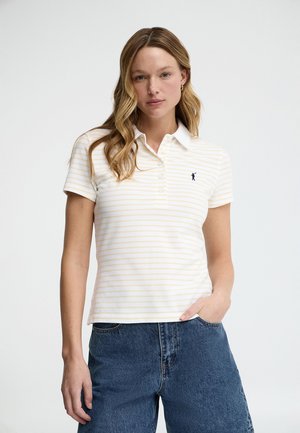 Femme aux cheveux blonds ondulés portant un polo rayé blanc et jaune clair et un jean en denim bleu, main dans la poche, fond neutre.