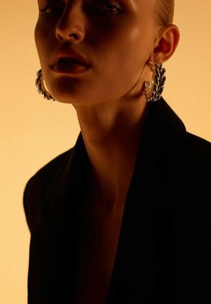 Velike srebrne obročeve uhane odlikuje zasukana, pletena zasnova. Oseba nosi črno blazer jakno na toplem, umirjenem ozadju.