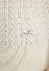 Crème kabelgebreide trui met een geribbelde zoom en een zilveren "JOOP!" label. Kenmerkt zich door een gestructureerd patroon, dat ingewikkelde weving toont.