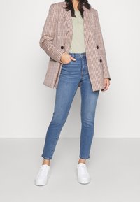 Rutig blazer i brunt och krämfärgat med dubbla knappar, kombinerad med åtsittande blå jeans och vita sneakers. Avslappnad design med modern passform.