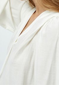 Bílá, texturovaná blůza s dlouhými rukávy a výstřihem do V, která má jemné vertikální pruhy a volný střih.