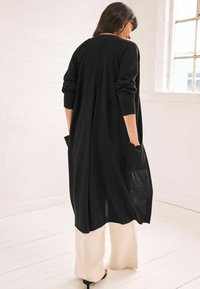 Lång svart cardigan i lätt stickad kvalitet, med ribbade ärmslut och sidofickor. Bärs över ljusfärgade, löst sittande byxor.
