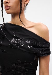 Melnbalta kleita bez plecu sīkumiem, ar ziedu sekvīru rotājumiem, teksturētu audumu un krītošu kakla izgriezumu, kas piešķir papildus detaļu.