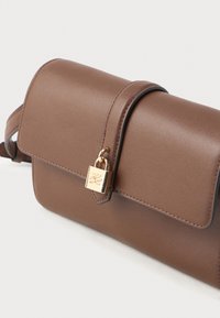 Brun skinn crossbody-veske med klaffelukking og liten gull hengelåsdetalj på forsiden mot en hvit bakgrunn.