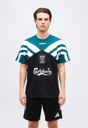 Young man wearing a black and teal Liverpool football jersey with Carlsberg and Adidas logos, standing against a plain light background.