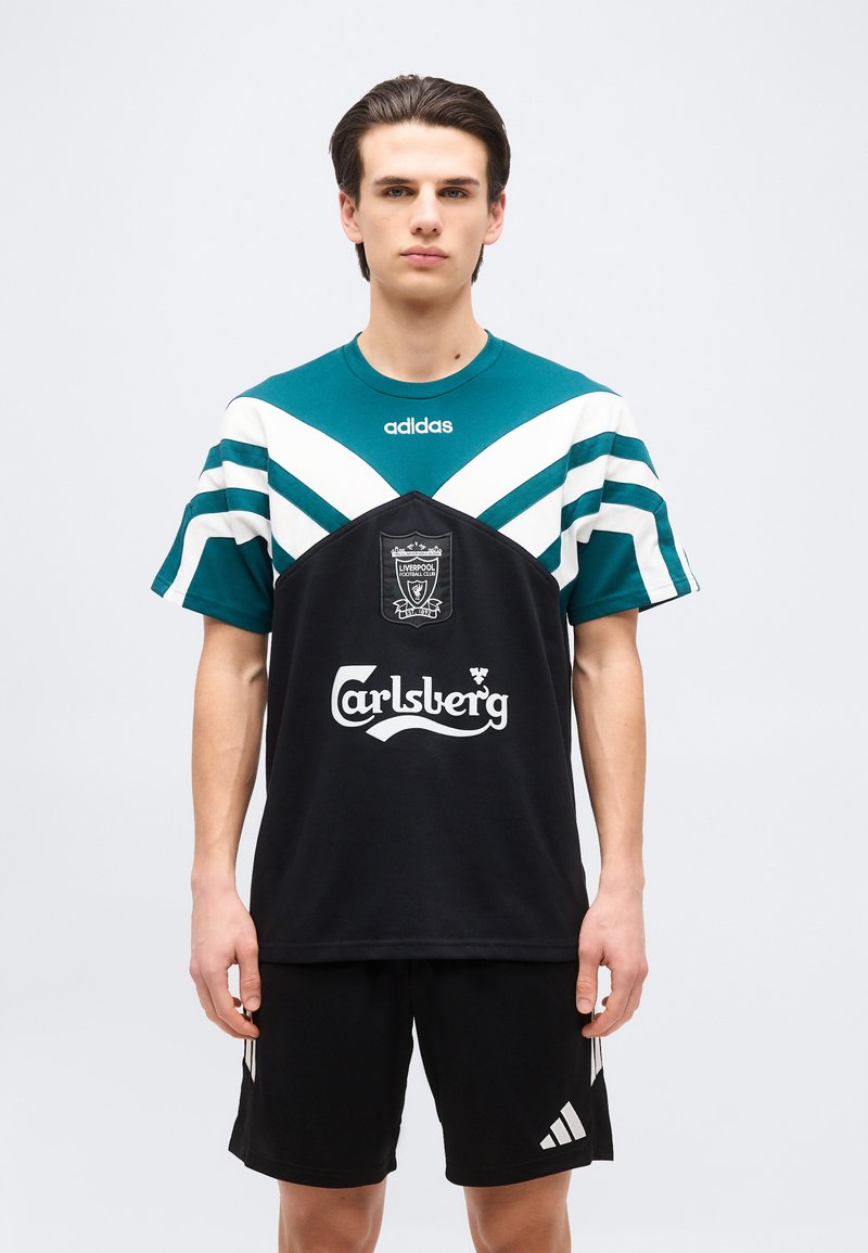 Jeune homme portant un maillot de football Liverpool noir et bleu sarcelle avec les logos Carlsberg et Adidas, debout devant un fond clair uni.