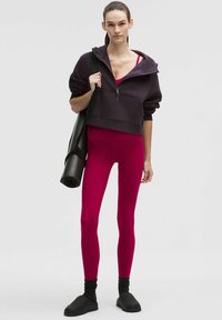 Fioletowa bluza z kapturem na zamek, fuksjowe legginsy, czarne skarpetki i wsuwane buty. Modelka trzyma zwiniętą matę do jogi. Gładkie tekstury i żywe kolory.