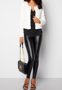 Hvid imiteret pelsjakke, sort top, skinnende sorte leggings, sort håndtaske med guldaccents og sorte hæle med stropper.