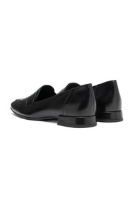Ett par svarta läder loafers utan snörning med låg klack, visade från en bakvinkel mot en vit bakgrund.