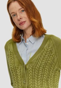 Zielona dziergana kardigan z wzorem w warkocze, dekoltem w kształcie litery V i zapięciem na guziki, noszony na jasnoniebieskiej koszuli w paski.