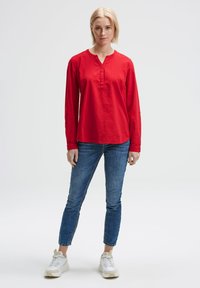 Rotes Langarmshirt mit Henley-Ausschnitt, aus weichem Stoff. Kombiniert mit blauen Slim-Jeans und weißen Sneakers, vor einem schlichten Hintergrund stehend.