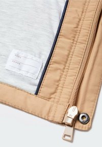 Giacca beige con chiusura a zip, con un interno in tessuto grigio e un'etichetta per i dettagli personali. Cuciture a vista e accessori in metallo.