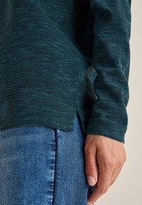 Ciemnozielony sweter o fakturowanym wzorze, z okrągłym dołem i bocznymi rozporkami. Noszony na niebieskich dżinsach, z widoczną ręką opartą na boku.