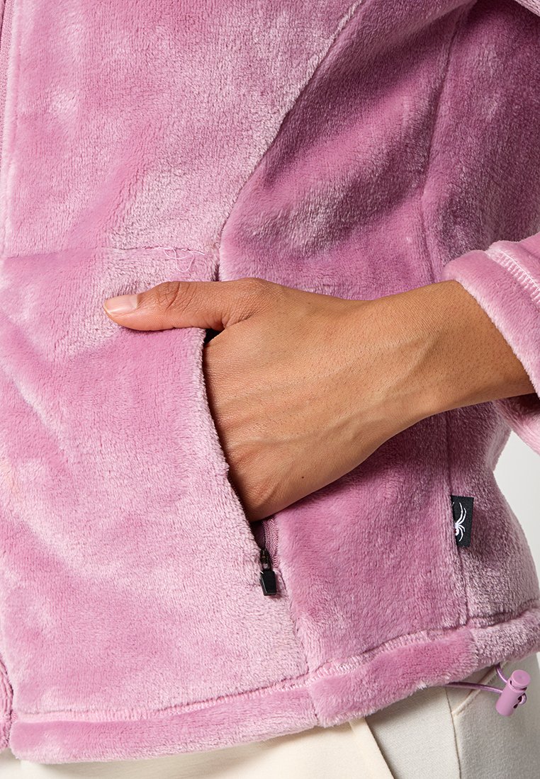 Różowa polarowa kurtka o miękkiej fakturze, z boczną kieszenią i małą metką marki z logo pająka blisko brzegu.