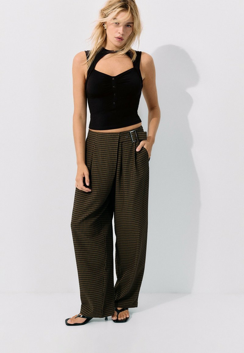 Femme debout portant un crop top noir sans manches et un pantalon large marron à carreaux taille haute avec des sandales sur fond blanc.