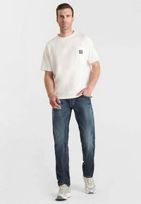 T-shirt bianca a maniche corte con taschino sul petto, abbinata a jeans denim azzurri. Il modello indossa sneaker grigie. Look semplice e casual.