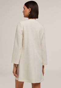 Rochie structurată de culoare crem, cu mâneci lungi, textură netedă și design simplu. Prezintă un spate curat, fără închizători vizibile.