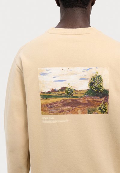 Krémová mikina se čtvercovým potiskem na zadní straně, který zobrazuje krajinu se stromy a kopci. Pod potiskem je text "*REWILDING."