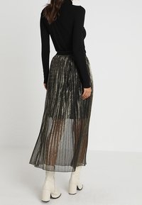 Jupe longue plissée en noir avec des accents métalliques dorés, en tissu transparent, et une taille ajustée, associée à un haut noir à manches longues.