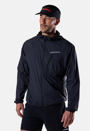 Man draagt een zwart Rossignol-jas met borstzak en een zwarte Rossignol-pet, kijkt zijwaarts tegen een effen lichte achtergrond.