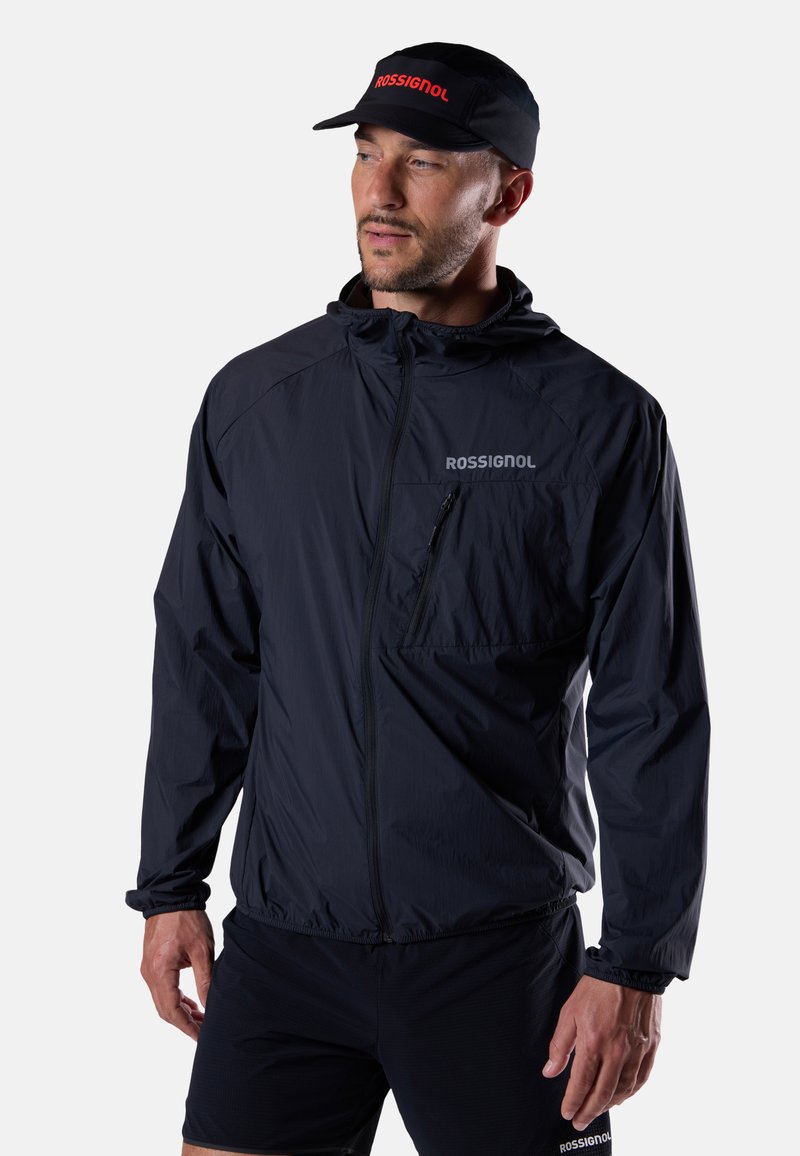 Mann trägt eine schwarze Rossignol-Jacke mit Brusttasche und schwarze Rossignol-Kappe, blickt seitlich vor einem schlichten hellen Hintergrund.