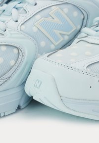 Baskets New Balance bleu clair et blanc avec des panneaux en tissu à pois et des logos "N" surélevés sur les côtés et les semelles.