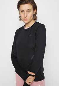 Crna dugorukava atletska majica s okruglim izrezom, teksturirana tkanina, suptilna oznaka branda i ružičasti detalj oko vrata.