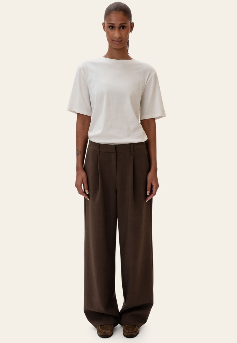 Femme debout portant une chemise blanche à manches courtes, un pantalon large marron et des chaussures marron sur un fond clair uni.