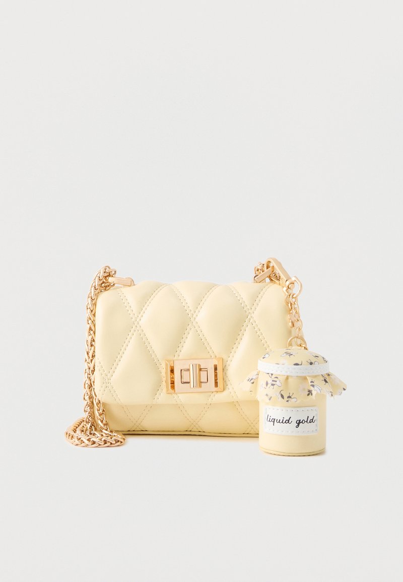 Petit sac matelassé jaune pâle avec bandoulière en chaîne dorée et bocal décoratif attaché étiqueté « or liquide » avec couverture en tissu floral.