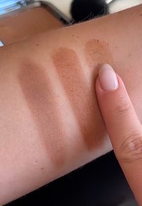 Trois échantillons de bronzer mat sur la peau : marron foncé à gauche, marron moyen au centre, marron clair à droite. Un doigt touche l'échantillon clair.