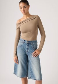 Ανοιχτό καφέ μακρυμάνικη μπλούζα με σχεδιασμό που αφήνει τους ώμους ακάλυπτους, συνδυασμένη με μπλε τζιν σόρτς culotte μεσαίου μήκους, με ξεθωριασμένο χρώμα και τσέπες.
