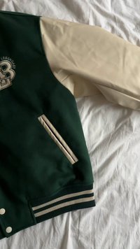 Green and beige varsity jacket with a round logo, ribbed cuffs, side pockets, and striped trim. Smooth fabric with contrasting sleeve colors.