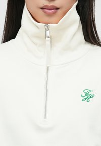 Baltas zip-up džemperis su aukšta apykakle, turintis žalią siuvinėtą logotipą. Lygus audinys su subtilia tekstūra, vidutinio svorio.