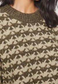 Detailní záběr na pletený svetr v zelené a krémové barvě s texturovaným geometrickým vzorem, který nosí osoba s dlouhými tmavými vlasy.