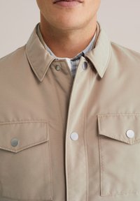Beige jasje van gladde stof met een puntige kraag, zilveren drukknoopsluitingen en twee voorzakken met kleppen.
