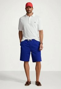 Mężczyzna w białej koszulce polo z krótkim rękawem, niebieskich szortach na sznurek, czerwonej czapce, brązowych butach żeglarskich i z zegarkiem na nadgarstku, stojący na jednolitym tle.