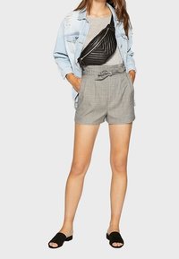 Donna che indossa una giacca di denim chiaro, un top grigio, pantaloni corti a quadri grigi a vita alta con cintura annodata, una borsa a tracolla nera e sandali neri senza chiusura.