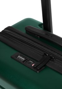 Valigia rigida verde con zip nera, meccanismo di bloccaggio e manico retrattile. Superficie texturizzata con accenti lucidi.