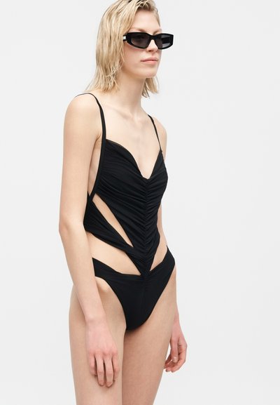 Γυναίκα με κοντά ξανθά μαλλιά, που φοράει μαύρο ολόσωμο μαγιό με σκισίματα και μαύρα ορθογώνια γυαλιά ηλίου, σε αφαιρούμενο φόντο.