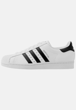 Baskets blanches Adidas Superstar avec trois bandes noires, embout shell toe et inscription « Superstar » sur le côté, vues de la face extérieure.