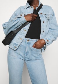 Hellblaues Denim-Jacket mit einem leicht übergroßen Schnitt, ausgestattet mit kupferfarbenen Knöpfen und zwei Brusttaschen, getragen mit einem schwarzen Oberteil und hochgeschnittenen Jeans.